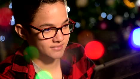 Joven,-el-niño-mira-a-la-tableta-tumbada-en-el-sofá.-En-el-fondo-luces-guirnaldas