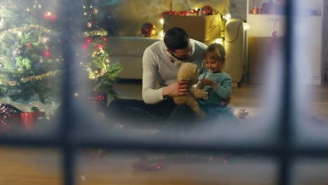 Mirando-a-través-de-Snowy-Window.-Sentado-bajo-Navidad-tres-padre-y-la-hija-juegan-con-el-oso-de-peluche.