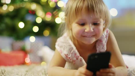 Niedliche-kleine-Mädchen-mit-Smartphone-in-der-Hand-liegt-auf-dem-Teppich-unter-dem-Weihnachtsbaum.