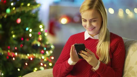 Hermosa-mujer-rubia-se-sienta-en-un-sofá-y-aplicaciones-Smartphone.-Árbol-de-Navidad-y-decoradas-con-luces-son-en-el-fondo.