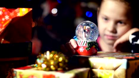 In-the-twilight-of-the-night,-among-the-gifts-in-bright-colorful-paper-packages,-a-pretty-little-blonde-girl-with-a-pink-bow-in-her-hair-and-a-beautiful-dress,-looks-carefully-and-admires-snow-globe.-toy-Christmas-ball-with-sparkles-and-a-snowman-inside