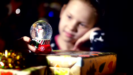 In-the-twilight-of-the-night,-among-the-gifts-in-bright-colorful-paper-packages,-a-pretty-little-blonde-girl-with-a-pink-bow-in-her-hair-and-a-beautiful-dress,-looks-carefully-and-admires-snow-globe.-toy-Christmas-ball-with-sparkles-and-a-snowman-inside