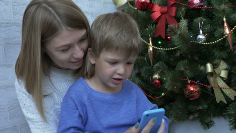 Fröhlicher-Junge-und-seine-Mutter,-Blick-auf-das-Smartphone-in-der-Nähe-von-Christmas-tree