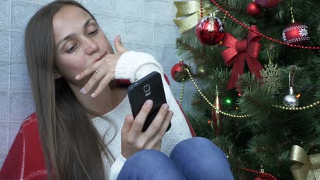 Frau-sitzt-auf-einer-Etage-mit-roten-Plaid-und-mit-Smartphone-neben-Weihnachtsbaum