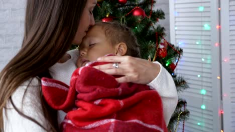 Joven-madre-con-su-pequeña-hija-dormida-en-las-manos-cerca-de-árbol-de-Navidad.