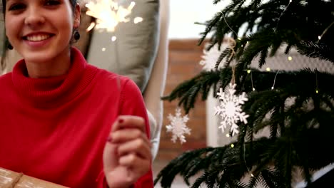 Smiling-friendly-attractive-young-woman-sitting-on-the-floor-by-the-Christmas-tree-celebrating-Xmas-with-sparklers-and-looking-in-the-camera