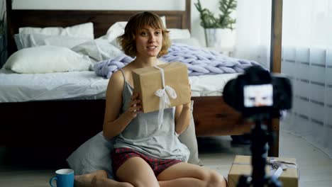 Attractive-cheerful-woman-sitting-near-bed-recording-video-blog-about-packing-gift-box-with-dslr-camera