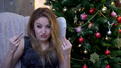 Stylish-young-woman-with-pink-lipstick-sitting-near-christmas-tree-and-showing-middle-finger