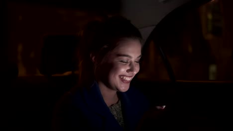 Niña-sonriente-feliz-montando-en-un-taxi-por-la-noche-sentado-en-el-asiento-trasero-y-usando-su-teléfono-inteligente-de-mensajes-de-texto.-Mujer-feliz-en-taxi.-En-la-noche.-Tiro-de-cámara-lenta