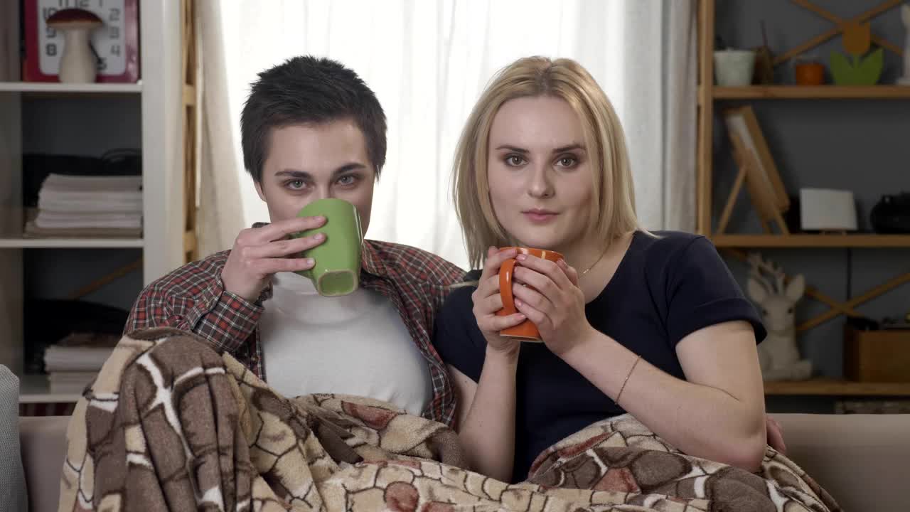 Two Young Lesbian Girls Are Sitting On The Couch, Covered With A Warm Blanket, Holding Cups In Their Hands, Drinking Dark Tea, Coffee, Cuddling, Looking At The Camera 60 Fps Free Stock Video Footage Download Clips Christmas