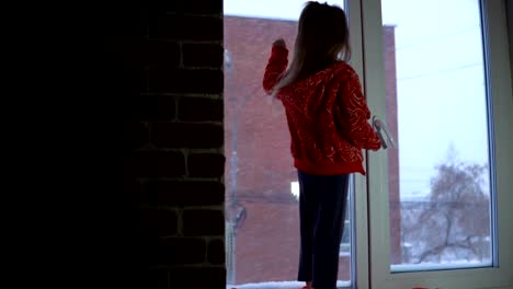 Little-cute-girl-standing-on-the-window-sill,-looking-out-on-a-snowy-cityscape.