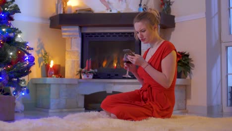 Young-girl-in-red-dress-sit-on-floor-near-Christmas-tree-and-using-her-phone