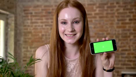 Young-fresh-ginger-girl-is-showing-green-screen-on-her-smartphone,-watching-at-camera,-communication-concept,-brick-background