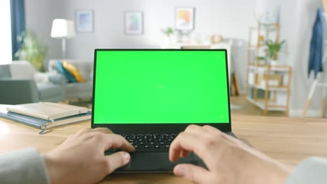 First-Person-Close-up-Shot-Man-Uses-Laptop-with-Green-Mock-up-Screen-While-Sitting-at-the-Desk-in-His-Cozy-Living-Room.
