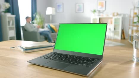 Modernen-Laptop-mit-grünen-Mock-up-Display-auf-dem-Schreibtisch-in-das-gemütliche-Wohnzimmer-stehen.-Mann-mit-Handy-sitzt-auf-einem-Stuhl,-Rest.