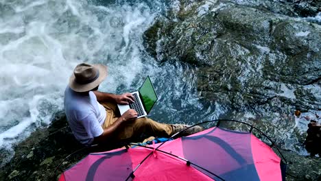 Junger-Mann-mit-green-Screen-Laptop-Computer-auf-einen-Wasserfall-und-Camping.-Reise--und-Freelance-Work-Konzept