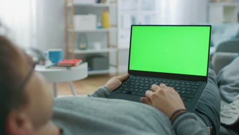 Mann-im-Hause-Verlegung-auf-eine-Couch-mit-grünen-Mock-Up-Screen-Laptop-Computer.-Mann-mit-Laptop-Gerät,-Internet-surfen,-Inhalte,-Videos-anschauen.