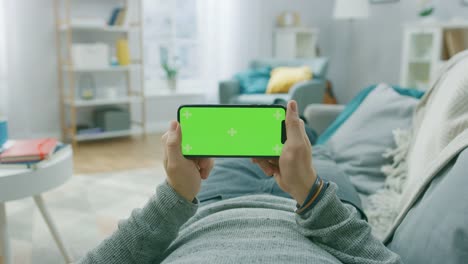 Mann-zu-Hause-hält-auf-einer-Couch-mit-Smartphone,-es-horizontal-im-Landscape-Modus.-Videospiele-spielen,-Videos-ansehen.-Bildschirm-hat-Tracking-Marker.-Point-Of-View-Kamera-gedreht.