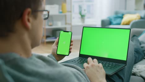 Young-Man-zu-Hause-arbeitet-auf-einem-Laptop-Computer-mit-Greenscreen-Mock-up-während-Holding-Smartphone-mit-Chroma-Key-Display.-Er-sitzt-auf-einer-Couch-in-seinem-gemütlichen-Wohnzimmer.-Über-die-Schulter-Kamera-Aufnahme