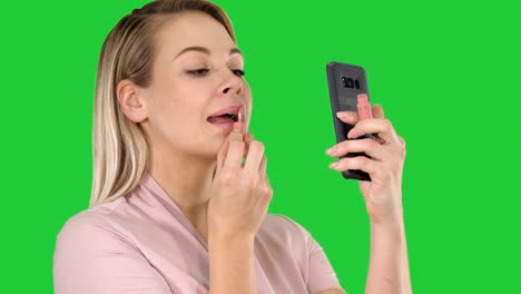 Beautiful-young-blonde-woman-which-is-painting-her-lips-with-red-lipstick-and-looking-in-the-phone-on-a-Green-Screen,-Chroma-Key