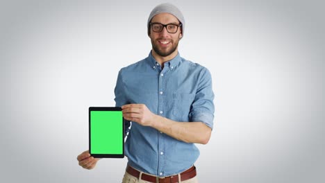 Mid-Shot-of-a-Fashionable-Young-Man-in-a-Hat-and-Glasses-Presenting-to-Us-Tablet-Computer-with-Isolated-Green-Screen.-Schuss-auf-einen-weißen-Hintergrund.