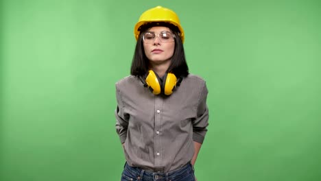 Mujer-joven-bonita-constructor-permanente-de-audífonos,-gafas-y-casco-con-las-manos-en-los-bolsillos-y-mirando-en-la-cámara,-fondo-clave-chroma