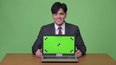 Young-handsome-Asian-businessman-showing-laptop-against-green-background