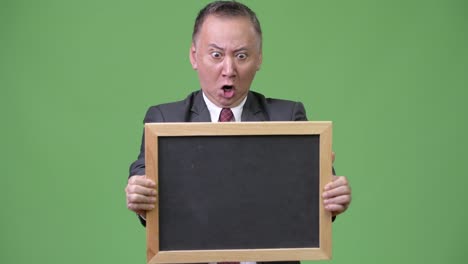 Mature-Japanese-businessman-holding-blackboard-against-green-background