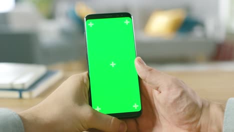 Primer-plano-de-un-hombre-que-sostiene-maqueta-verde-pantalla-Smartphone-en-sus-manos.-Utilizando-dispositivo-inalámbrico-para-navegar-por-Internet.-En-el-fondo,-acogedora-sala-de-estar-o-Home-Office.