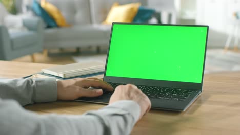 Close-up-of-a-Man-Uses-Laptop-with-Green-Mock-up-Screen-While-Sitting-at-the-Desk-in-His-Cozy-Living-Room.