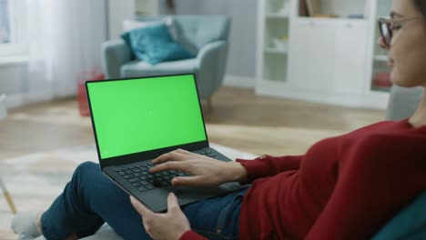 Junge-Frau-zu-Hause-arbeitet-an-einem-Laptop-Computer-mit-grünen-Mock-up-Bildschirm.-Sie-sitzt-auf-einer-Couch-in-seinem-gemütlichen-Wohnzimmer.-Über-die-Schulter-Kamera-Aufnahme