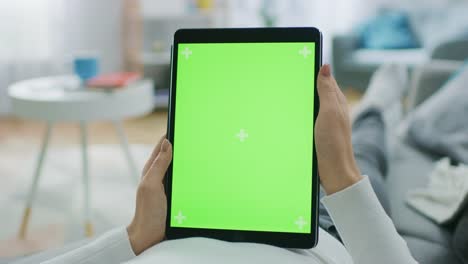 Young-Woman-at-Home-Laying-on-a-Couch-Using-with-Green-Mock-up-Screen-Tablet-Computer-in-Vertical-Portrait-Mode.-Man-Using-Touchscreen-Device,-Browsing-Internet,-Watching-Content,-Videos.-POV.