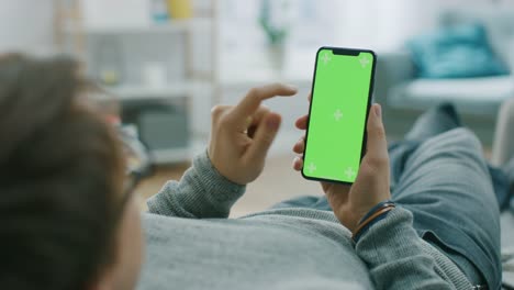 Mann-zu-Hause-auf-der-Couch-mit-Smartphone-mit-Mock-up-Greenscreen,-tun,-streichen,-scrollen-Gesten-liegen.-Mann-mit-Handy,-soziale-Netzwerke-im-Internet-surfen.