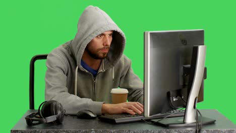 Tiro-medio-de-un-Hacker-con-Hoodie-sentado-en-su-computadora-de-escritorio.-El-fondo-de-pantalla-verde.