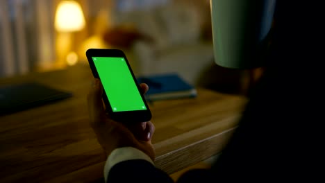 At-Home-Man-Sits-at-His-Desk-and-Uses-Smartphone-with-Green-Screen-on-It-Er-Drinkt-aus-einem-Cup.-Seine-Wohnung-ist-in-Gelben-Farben-gehalten-und-ist-warm.