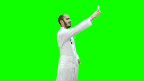 Sonriente-médico-de-bata-blanca-con-estetoscopio-tomando-selfie-en-su-teléfono-en-una-pantalla-verde-Chroma-Key