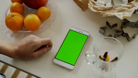 Closeup-of-woman's-hand-browsing-smartphone-with-green-screen-on-kitchen-table-at-home