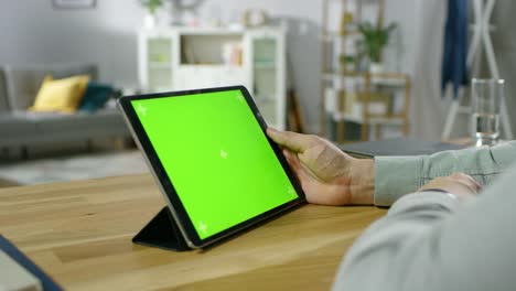 Über-die-Schulter-geschossen-von-einem-Mann-Betrieb-und-beobachtete-grüne-Mock-up-Bildschirm-Digital-Tablet-Computer-sitzend-am-Schreibtisch.-Im-gemütlichen-Wohnzimmer-Hintergrund.