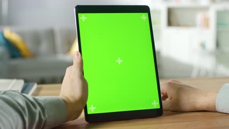 Close-up-of-Man-Using-Hand-Gestures-on-Green-Mock-up-Screen-Digital-Tablet-Computer-in-Portrait-Mode-while-Sitting-at-His-Desk.-In-the-Background-Cozy-Living-Room.