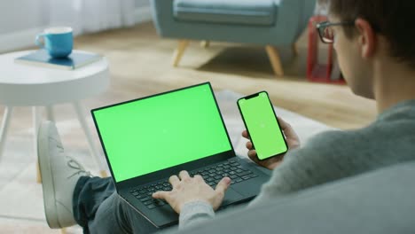 Young-Man-zu-Hause-arbeitet-auf-einem-Laptop-Computer-mit-Greenscreen-Mock-up-während-Holding-Smartphone-mit-Chroma-Key-Display.-Er-sitzt-auf-einer-Couch-in-seinem-gemütlichen-Wohnzimmer.-Über-die-Schulter-Kamera-Aufnahme