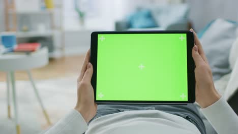 Young-Woman-at-Home-Laying-on-a-Couch-Using-with-Green-Mock-up-Screen-Tablet-Computer-in-Horizontal-Landscape-Mode.-Girl-Using-Touchscreen-Device,-Browsing-Internet,-Watching-Content,-Videos,-Blogs.