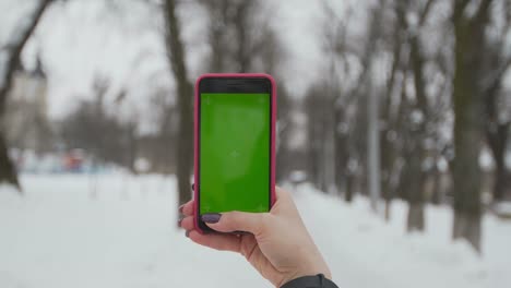Young-woman-types-and-slides-on-her-smartphone-in-the-winter-park.-Cellphone-with-chroma-key-screen---green-screen.Close-up.-Tracking-motion.