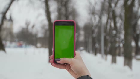 Junge-Frau-Typen-und-Folien-auf-ihrem-Smartphone-im-Winter-Park.-Handy-mit-Chroma-Key---grüner-Bildschirm.-Hautnah.-Verfolgen-von-Bewegung.