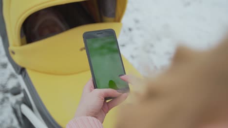 Young-woman-types-and-slides-on-her-smartphone-in-the-winter-park.-Cellphone-with-chroma-key-screen---green-screen.Close-up.-Tracking-motion.