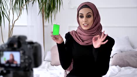 Young-beautiful-Indian-girl-in-hijab-recommends-an-application-on-a-smartphone,-smiling,-talking-at-the-camera,-chroma-key,-green-screen-concept.-50-fps