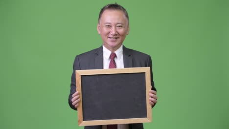 Mature-Japanese-businessman-holding-blackboard-against-green-background