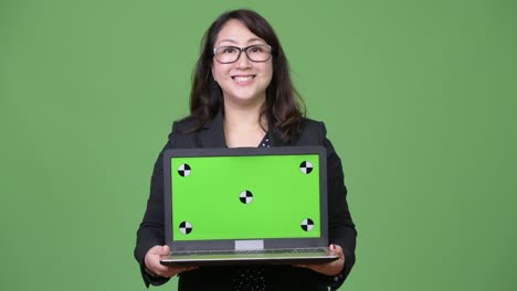 Mature-beautiful-Asian-businesswoman-showing-laptop-against-green-background