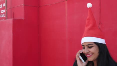 Handheld-shot-of-Indian-woman-with-Santa's-hat-talking-on-mobile-cell-phone,-cheerful-and-excited,-with-red-background