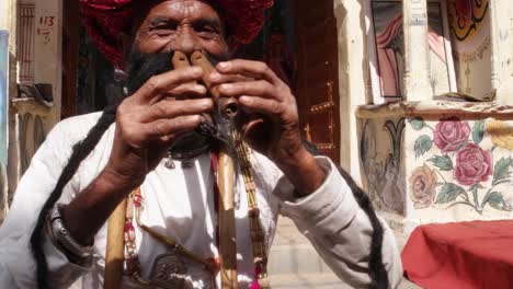 Mano-Rajasthani-ancianos-hombre-toca-la-flauta-con-la-nariz-delante-de-un-arco-del-templo-pintado,-con-gran-bigote-con-atuendos-tradicionales