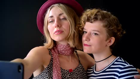 Two-charming-stylish-young-women-with-short-hair-embracing-and-taking-selfie-,-isolated-on-black-background